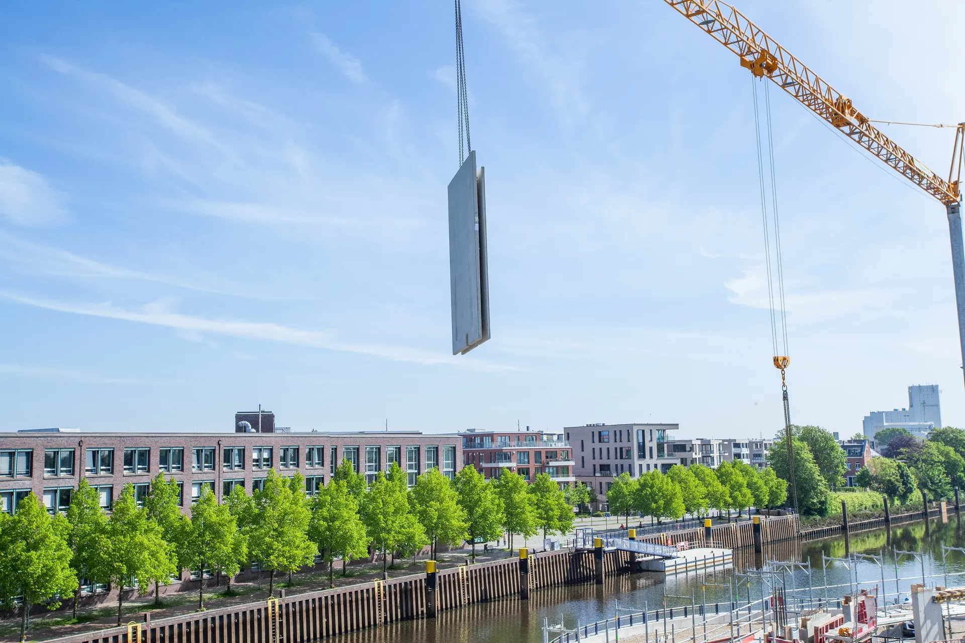 Eine große Betonfertigteilwand wird mit einem Kran über einer Baustelle angehoben. Im Vordergrund verläuft ein Fluss mit Spundwänden, dahinter stehen moderne Gebäude und eine Allee mit grünen Bäumen. Der Himmel ist blau und klar.

                                  